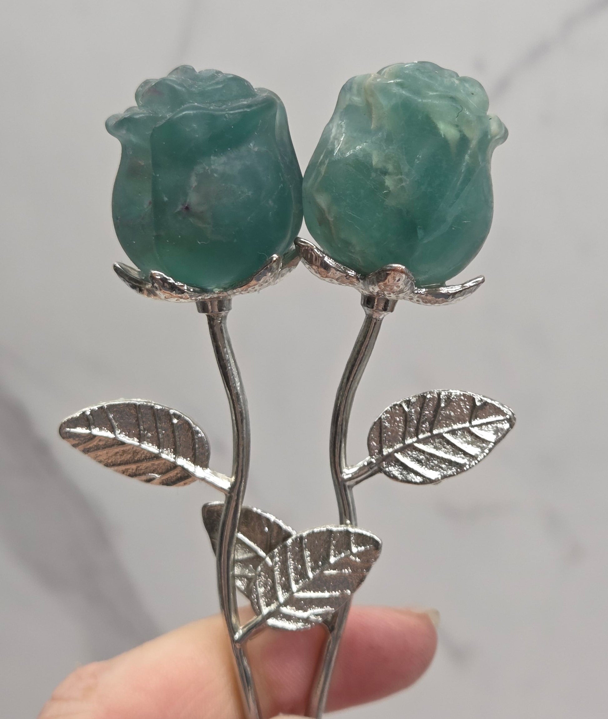 A close-up of a hand holding a silver-toned hair claw clip designed as two green gemstone roses with leaves, set against a light gray marble background.