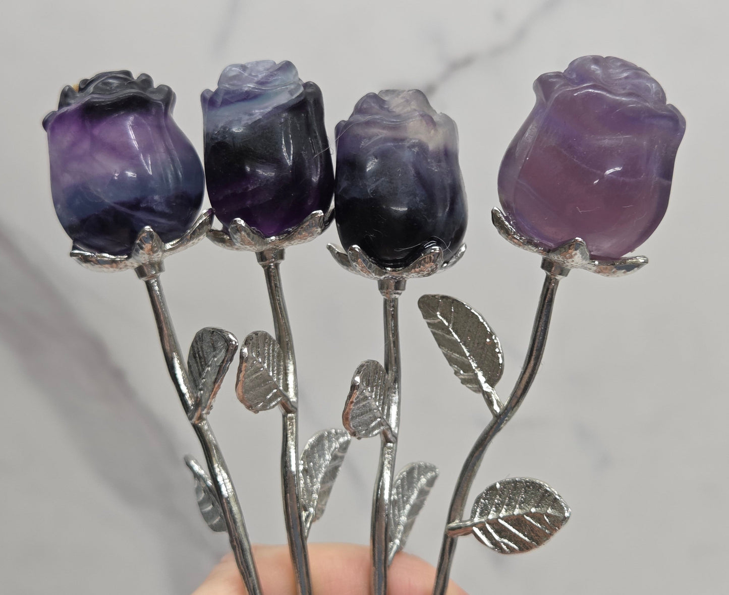 Four silver rose-shaped hair sticks with purple fluorite flower buds, held by a hand against a light gray marble background.