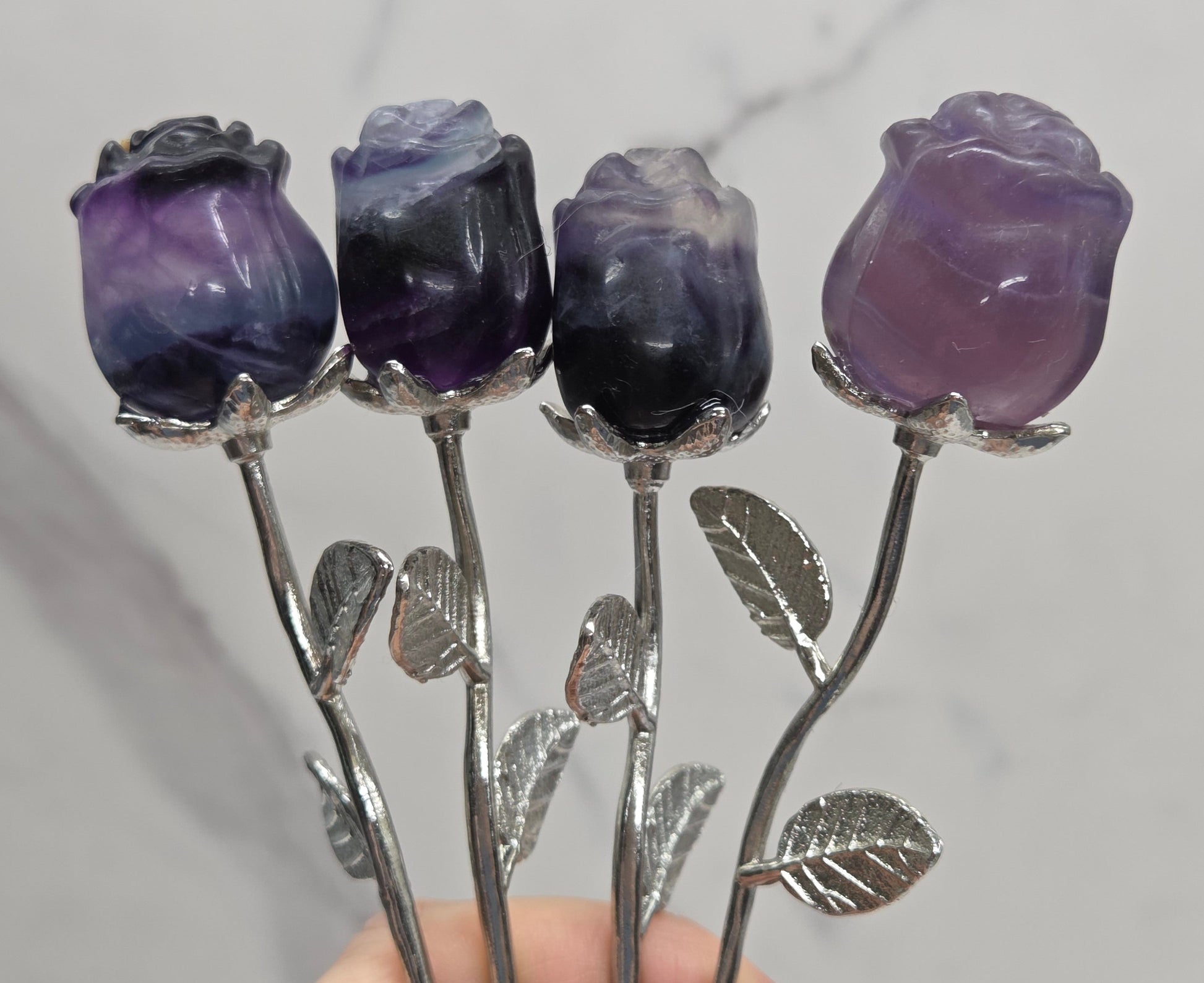 Four silver rose-shaped hair sticks with purple fluorite flower buds, held by a hand against a light gray marble background.