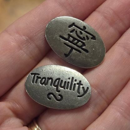 Two small oval silver-toned metal tags engraved with symbols and the word 'tranquility' are held in a person's hand over a wooden surface.