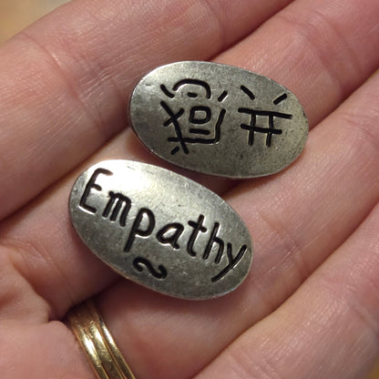 Two oval silver-toned metal pendants with engraved text, one with Chinese characters and the other reading 'Empathy co.', held in a person's hand over a wooden surface.