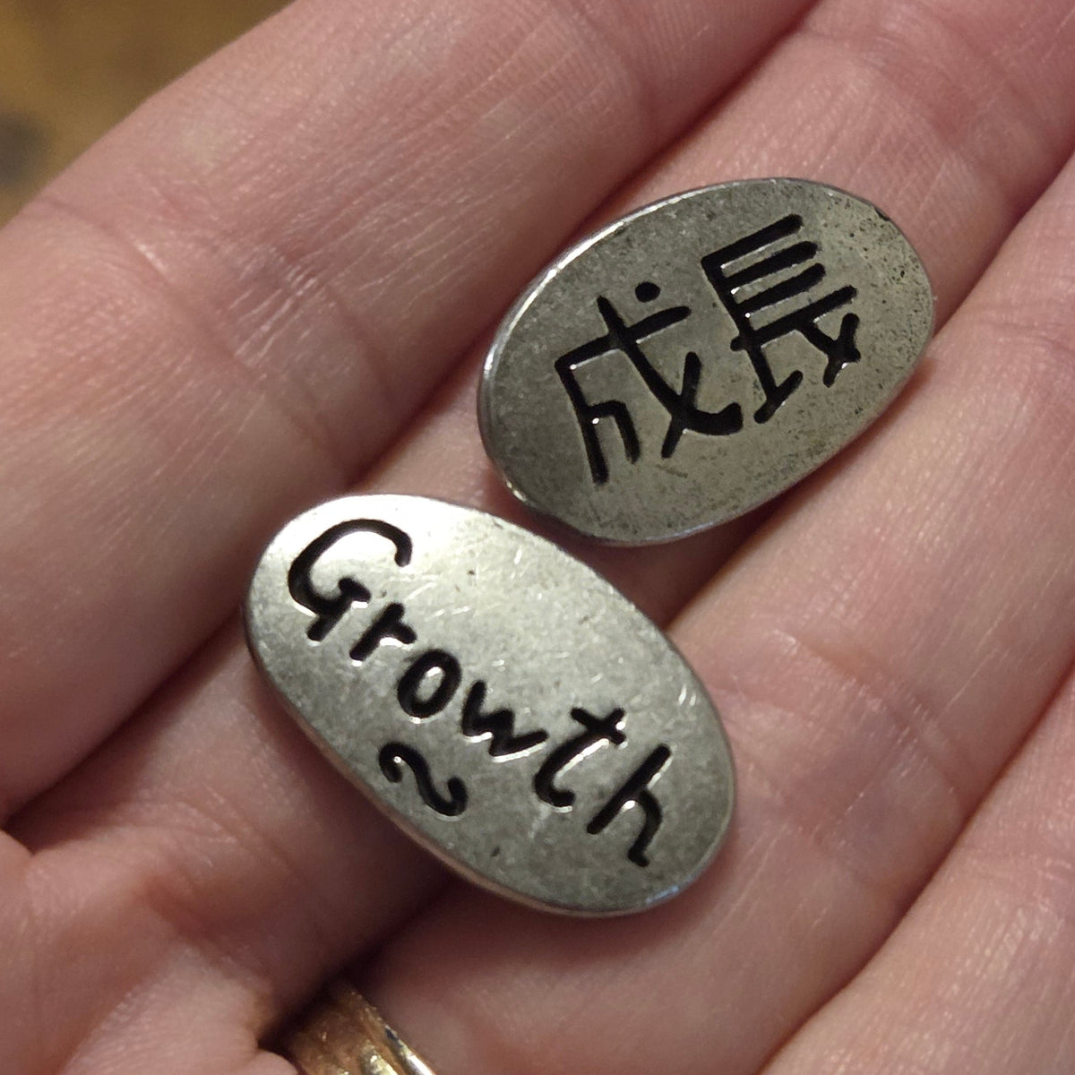 Two small oval silver metal tags engraved with Chinese characters and the word 'Growth' are held in a person's hand over a wooden surface.
