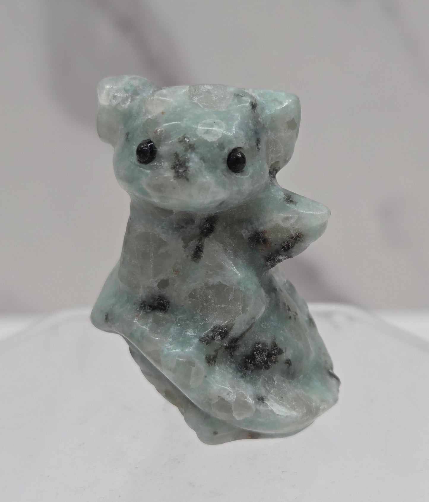 A small carved stone figurine of a dragon made from light green and white marbled stone with black speckles, displayed on a clear acrylic stand against a white marble background.
