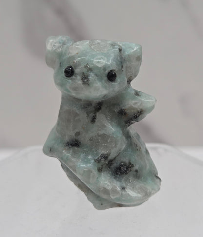 A small carved stone figurine of a dragon made from light green and white marbled stone with black speckles, displayed on a clear acrylic stand against a white marble background.