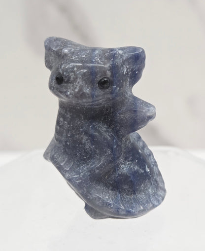 A small carved stone figurine of a dragon, made from a dark blue-gray material with visible grain, displayed on a clear acrylic stand against a light marble background.