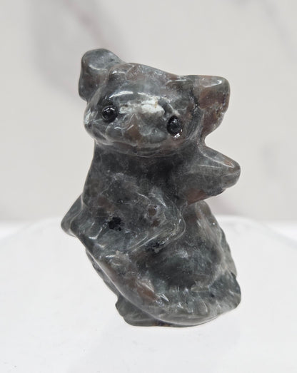A small carved figurine of a dragon, made from dark gray stone with natural marbling, shown on a clear acrylic stand against a white marble background.