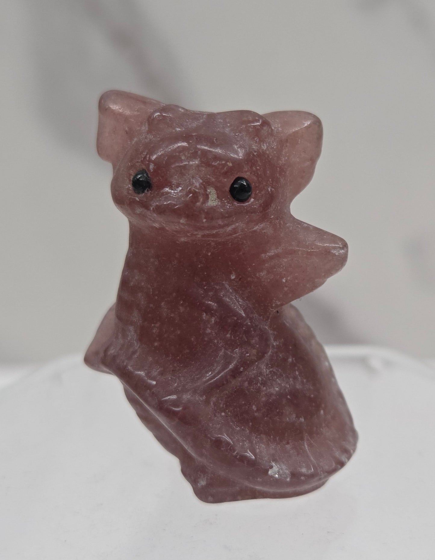 A small carved figurine of a dragon in dusty rose quartz, featuring small black bead eyes, displayed on a clear acrylic stand against a light gray marble-patterned background.