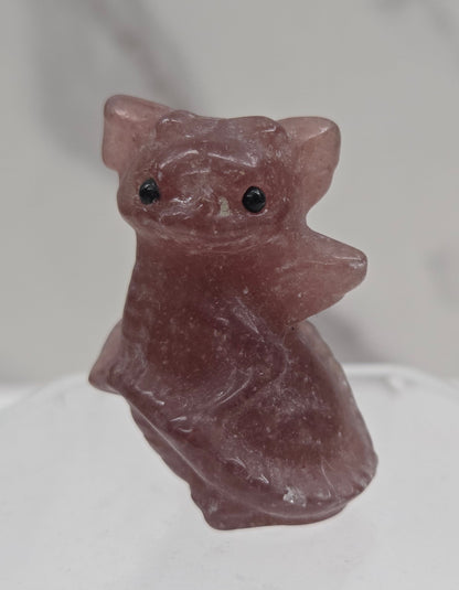 A small carved figurine of a dragon in dusty rose quartz, featuring small black bead eyes, displayed on a clear acrylic stand against a light gray marble-patterned background.