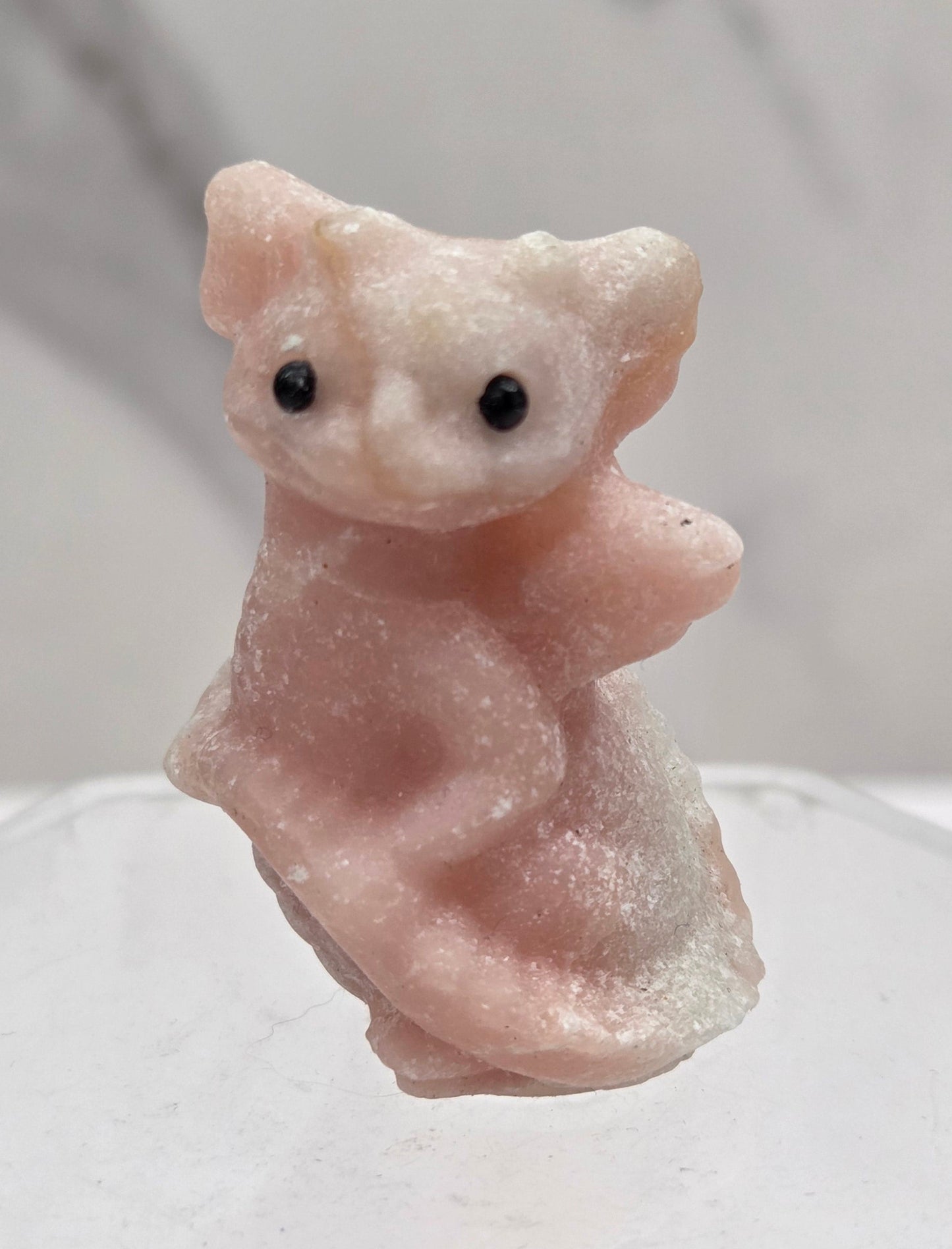 A small carved pale pink stone figurine with black eyes, shown close up against a light gray marble-like background and a translucent white hexagonal object on the right.