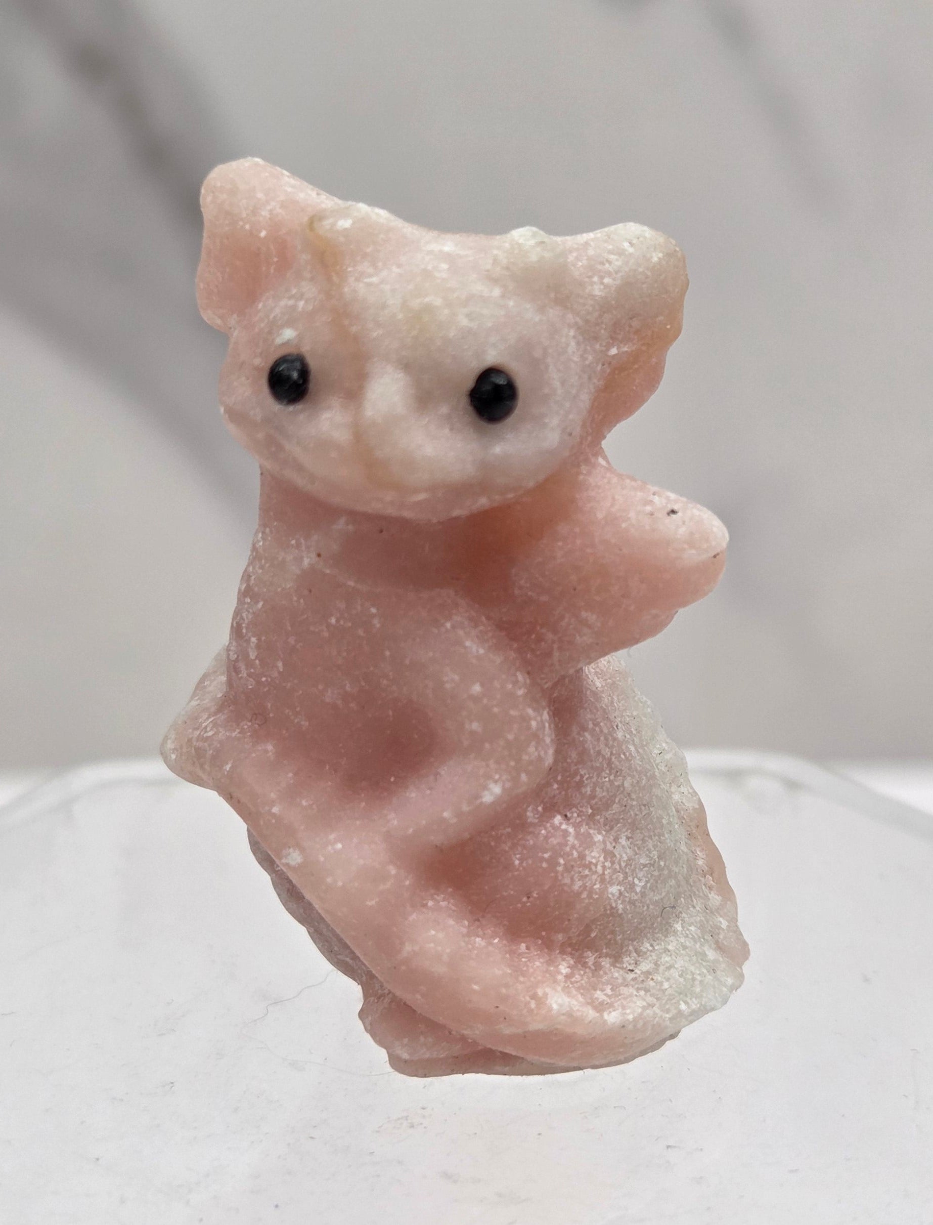 A small carved pale pink stone figurine with black eyes, shown close up against a light gray marble-like background and a translucent white hexagonal object on the right.
