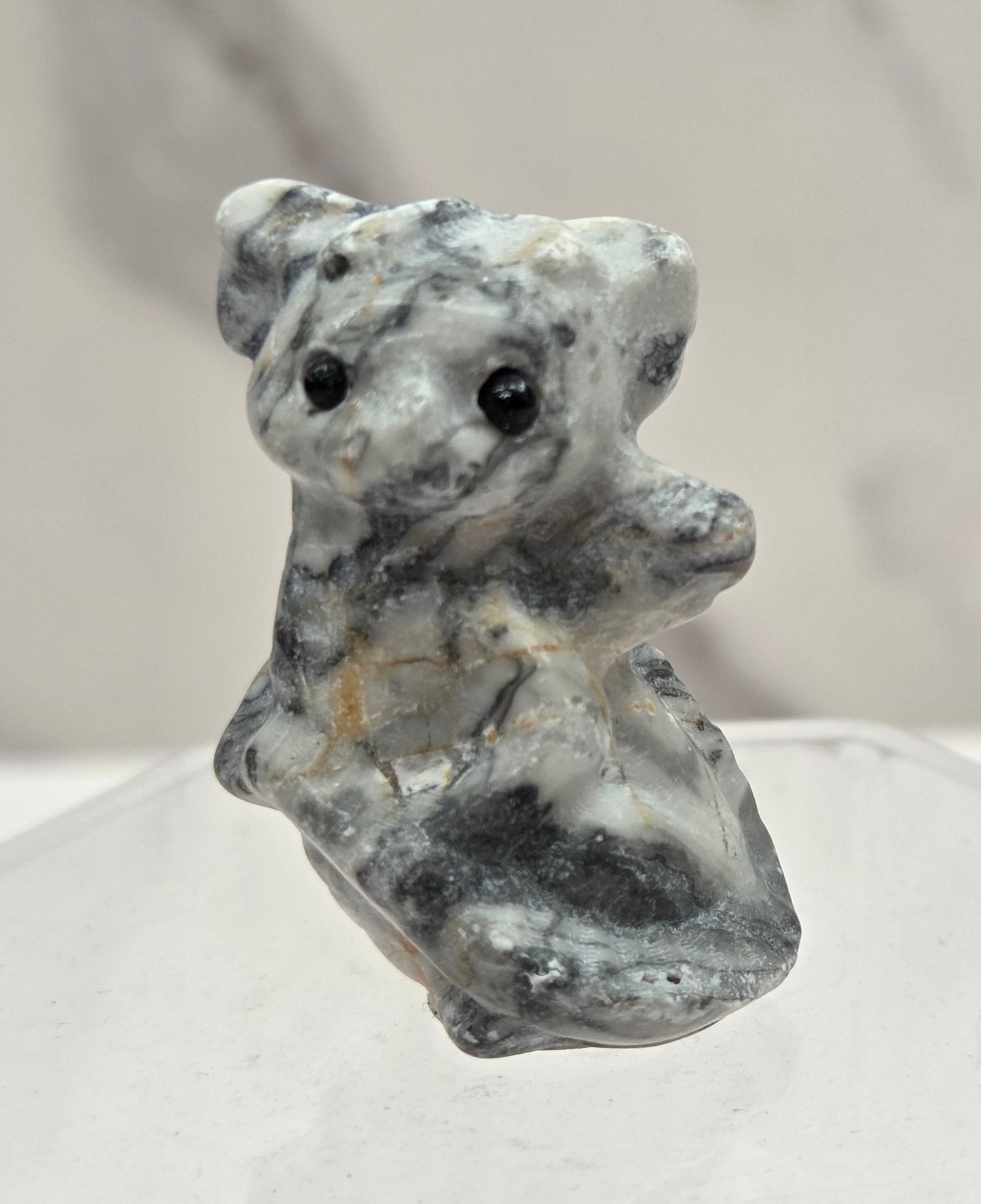 A small carved figurine of a dragon made from gray and white marbled stone, displayed on a clear acrylic stand against a light gray marble background.