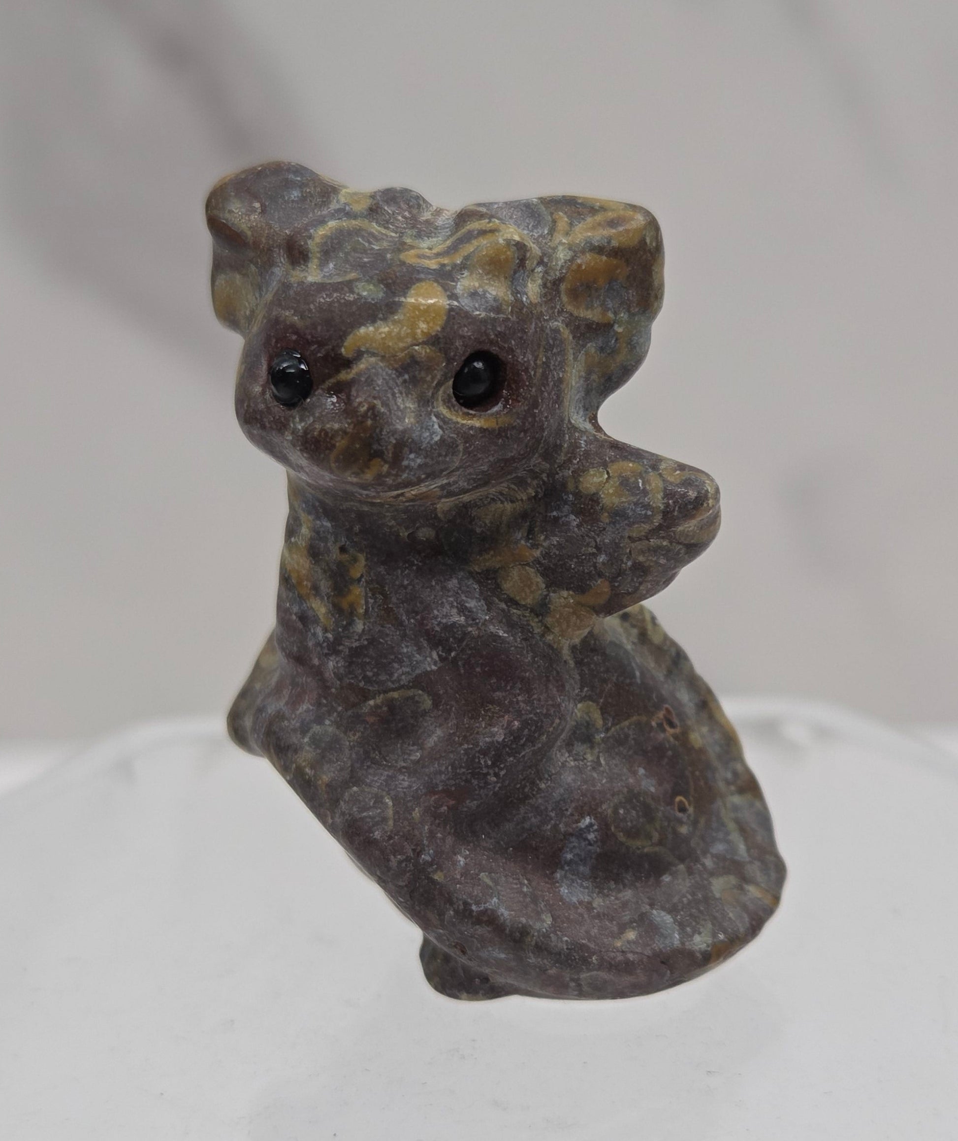 A small carved figurine of a dragon, made from dark gray stone with subtle gold and greenish patina details, shown against a white marble-like background.