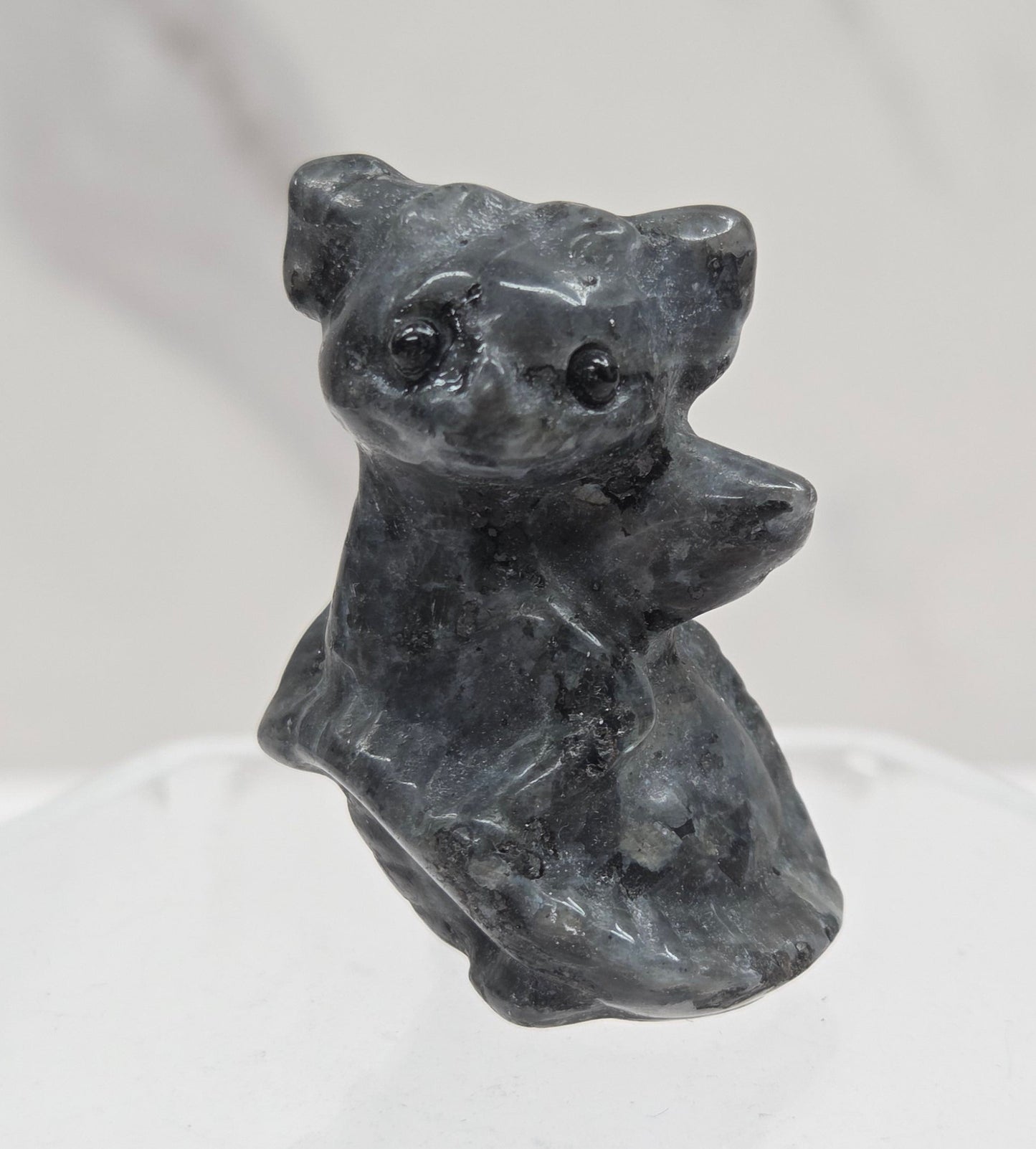 A small sculptural dog figurine in dark charcoal gray stone, mounted on a clear acrylic hexagonal display stand against a light gray marble-textured background.