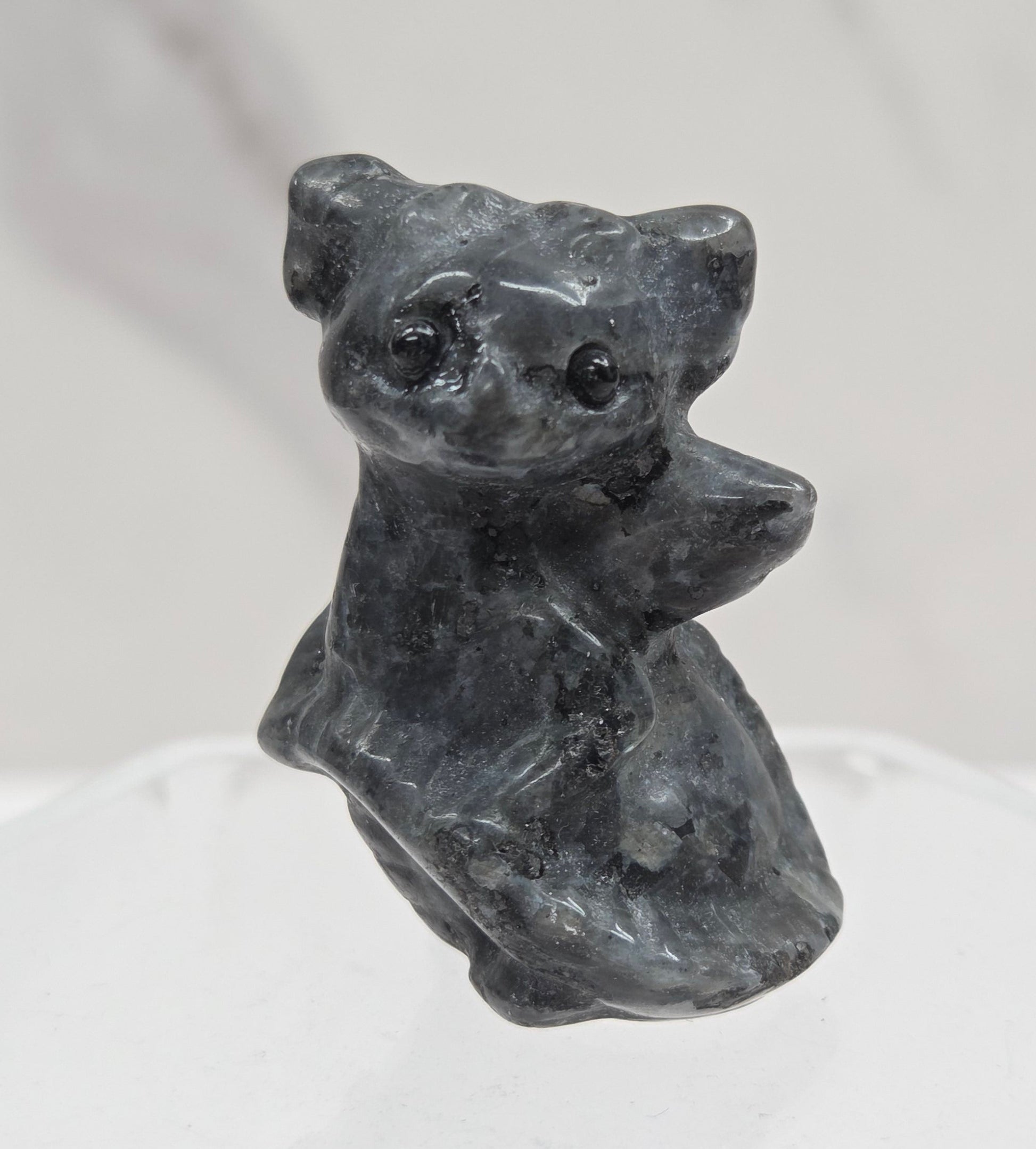 A small sculptural dog figurine in dark charcoal gray stone, mounted on a clear acrylic hexagonal display stand against a light gray marble-textured background.