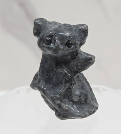 A small sculptural dog figurine in dark charcoal gray stone, mounted on a clear acrylic hexagonal display stand against a light gray marble-textured background.