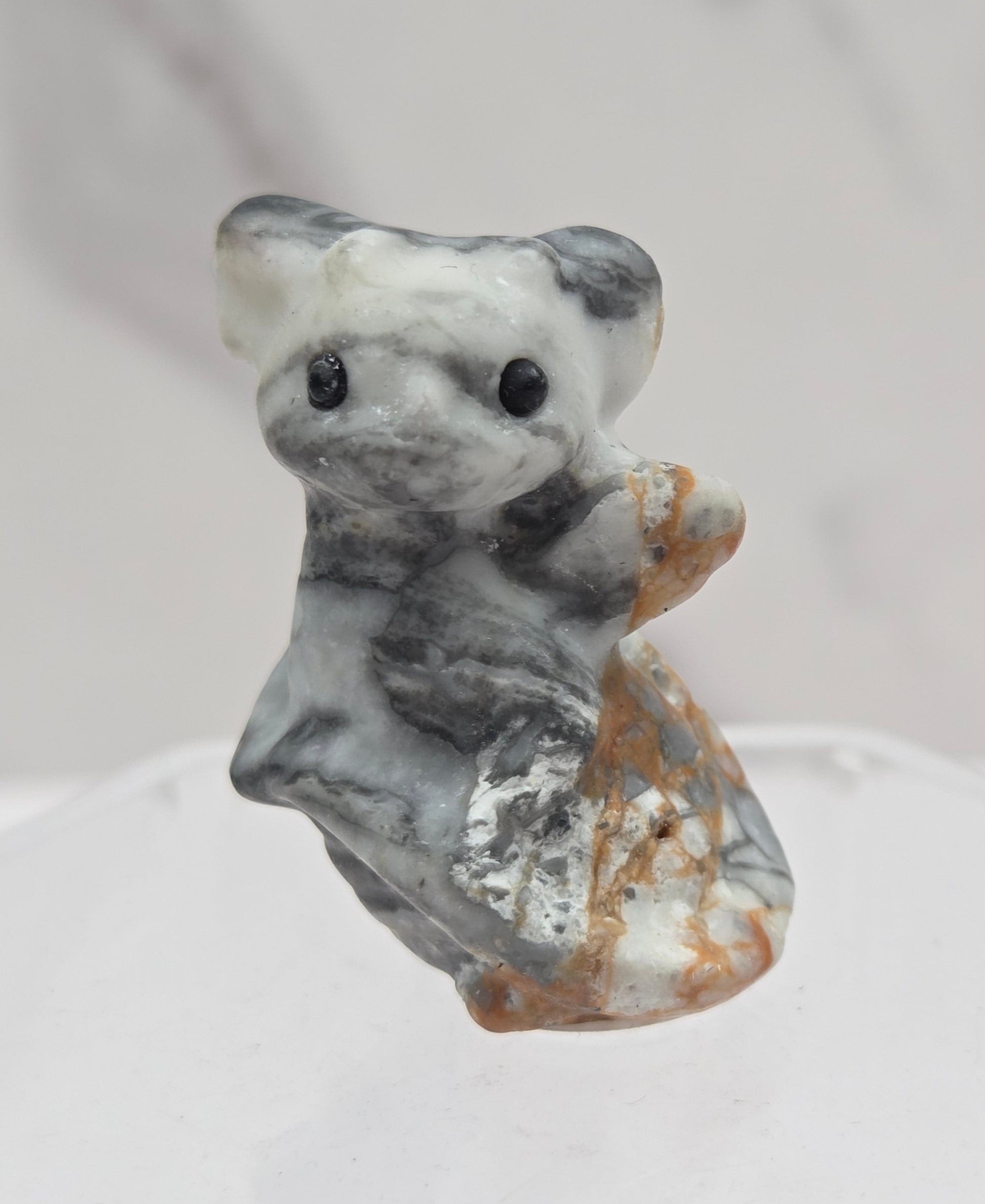 A small carved stone figurine of a gray and white dragon with brown accents, mounted on a clear hexagonal display stand against a light gray marble-textured background.