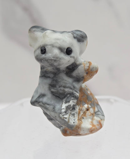 A small carved stone figurine of a gray and white dragon with brown accents, mounted on a clear hexagonal display stand against a light gray marble-textured background.