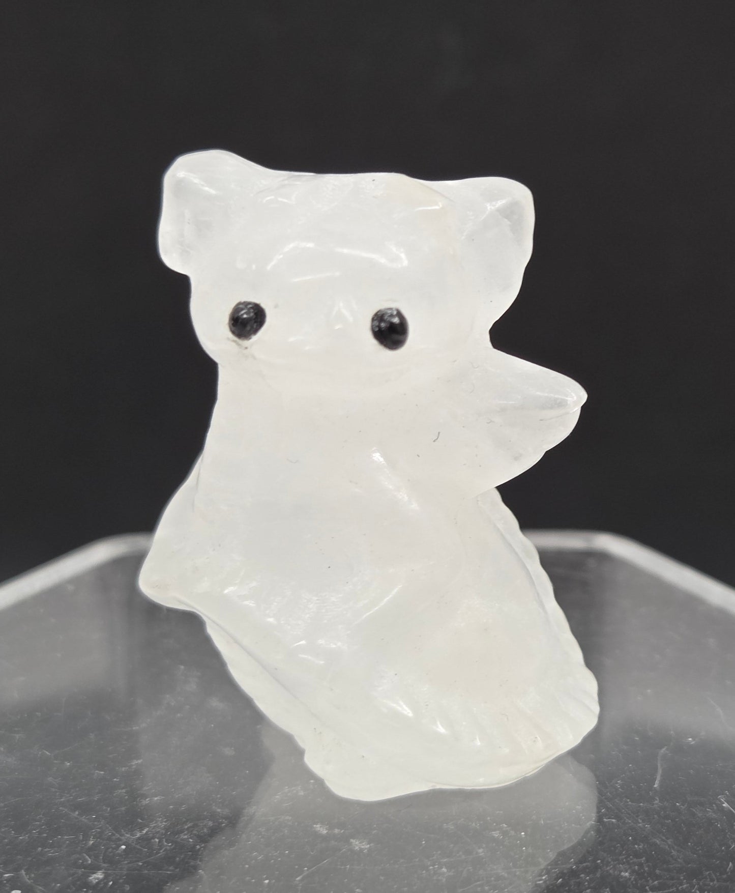 A small carved white animal figurine with black eyes, shown beside a clear acrylic display stand against a dark background.