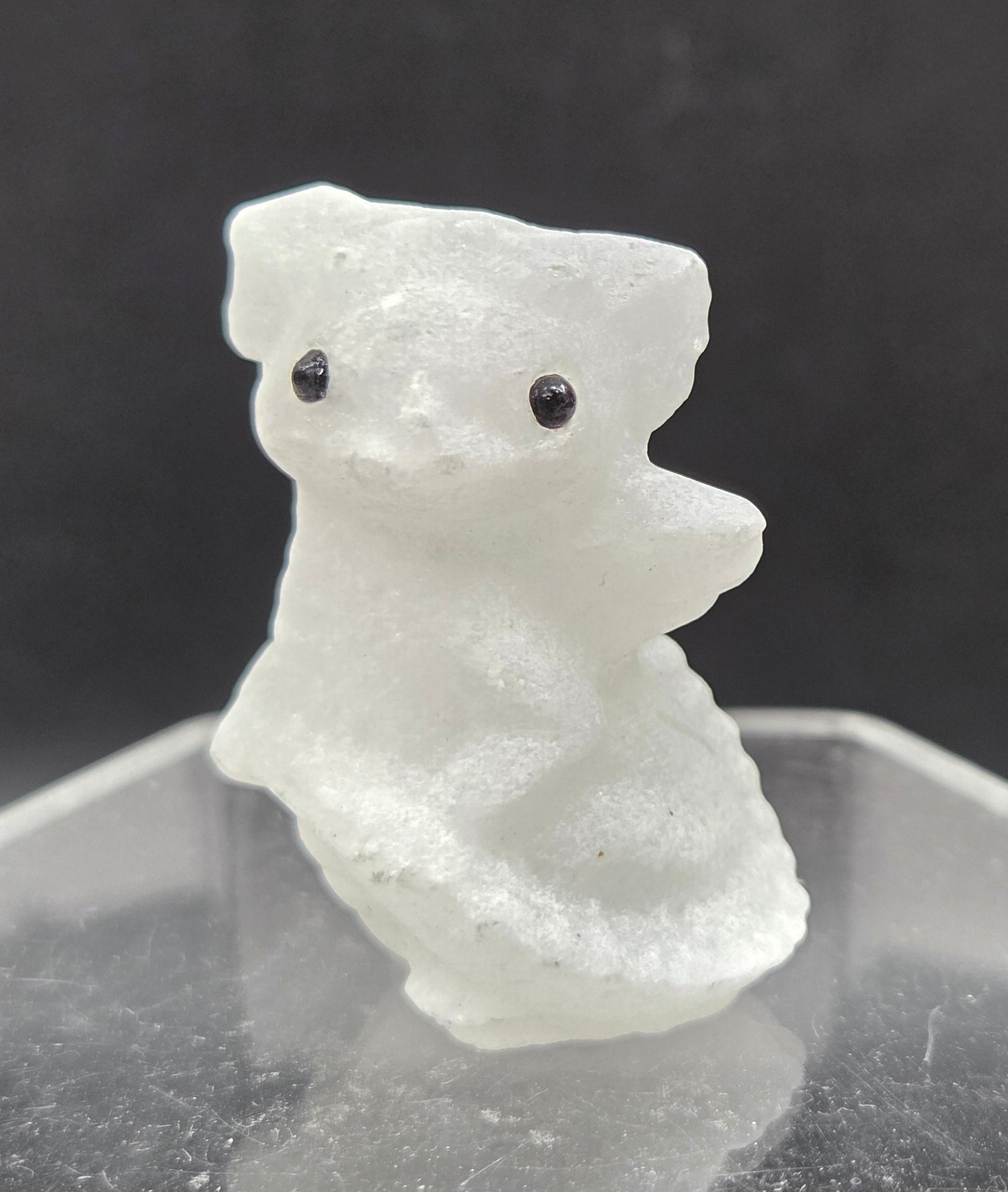 A small translucent white carved figurine resembling a dragon with two black eyes, mounted against a clear geometric acrylic stand on a dark gray background.