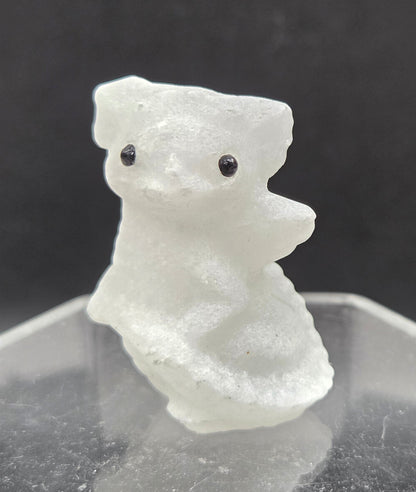 A small translucent white carved figurine resembling a dragon with two black eyes, mounted against a clear geometric acrylic stand on a dark gray background.