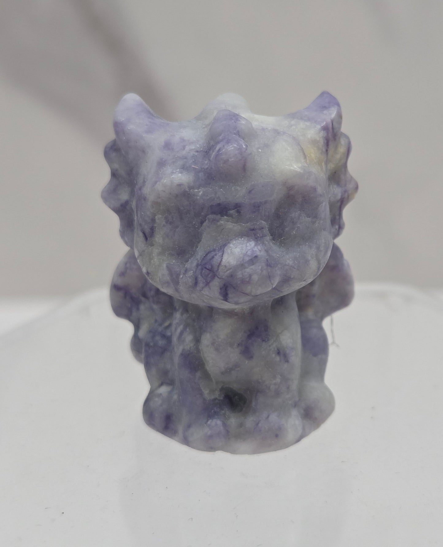 A small sculptural figurine of a stylized animal head, likely a stone or resin creature, in marbled lavender, purple, and white tones, mounted on a clear geometric display stand against a light gray background.