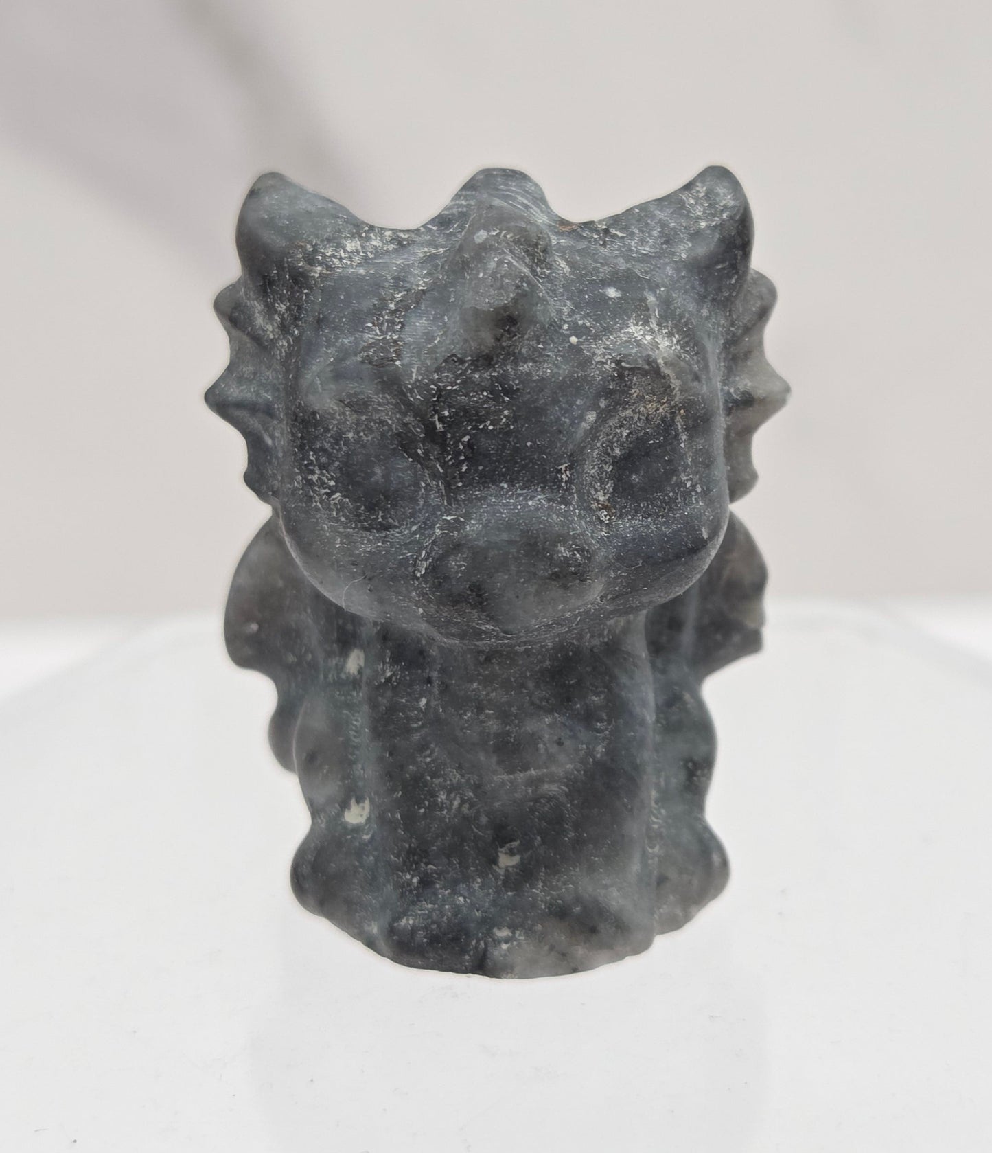 A small dark gray stone or resin figurine with a textured, organic shape, mounted on a clear acrylic stand against a white marble-patterned background.