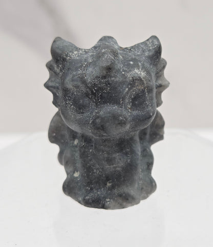 A small dark gray stone or resin figurine with a textured, organic shape, mounted on a clear acrylic stand against a white marble-patterned background.