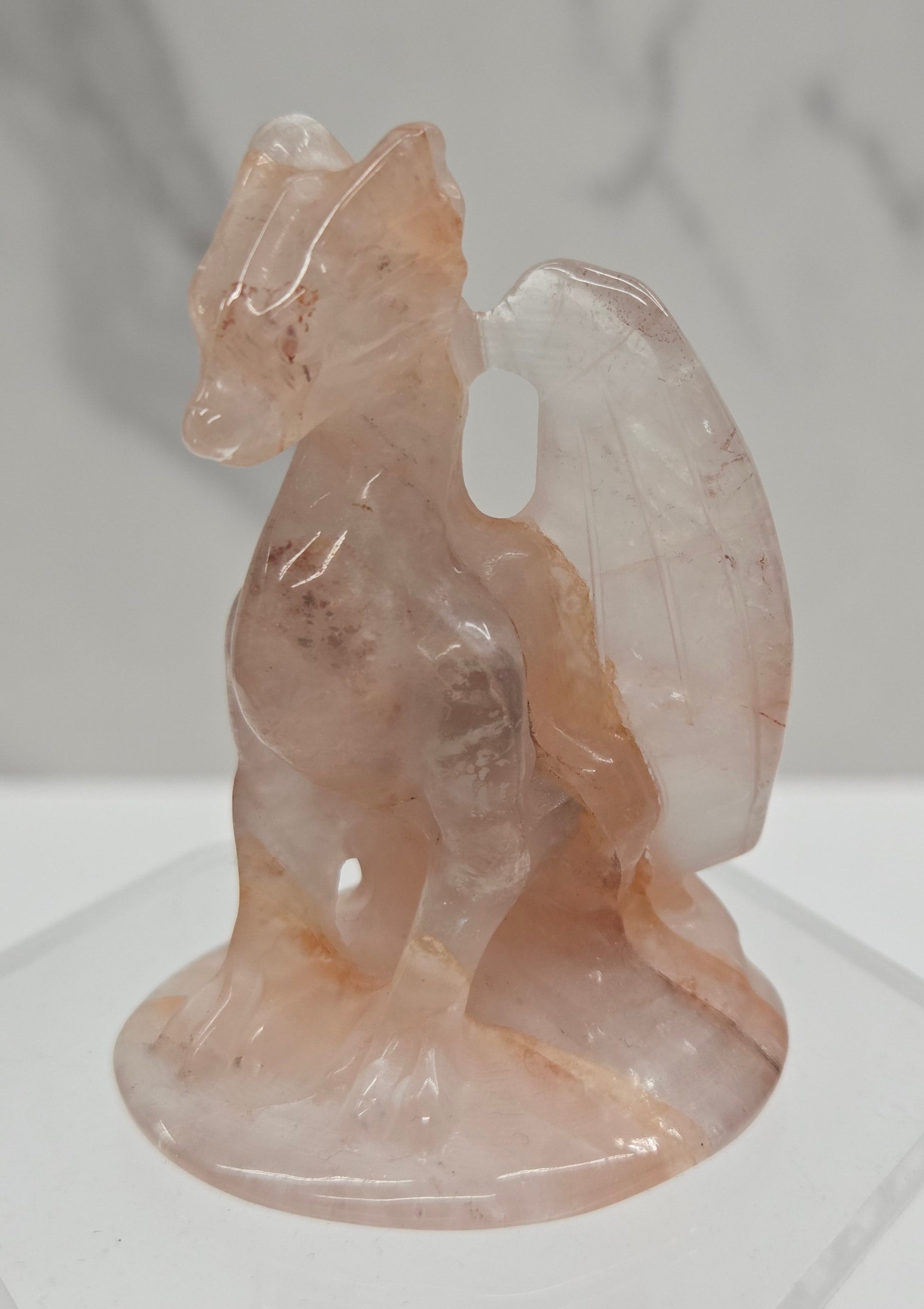 A translucent pale pink carved stone sculpture mounted on a clear acrylic stand against a white and gray marble-patterned background.