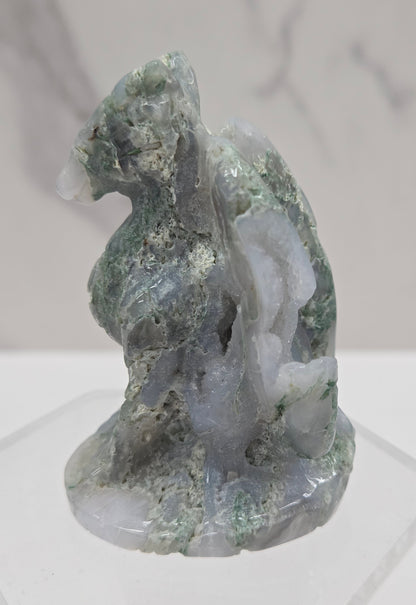 A carved dragon, featuring shades of green, gray, and white, mounted on a clear hexagonal display stand against a light gray background.
