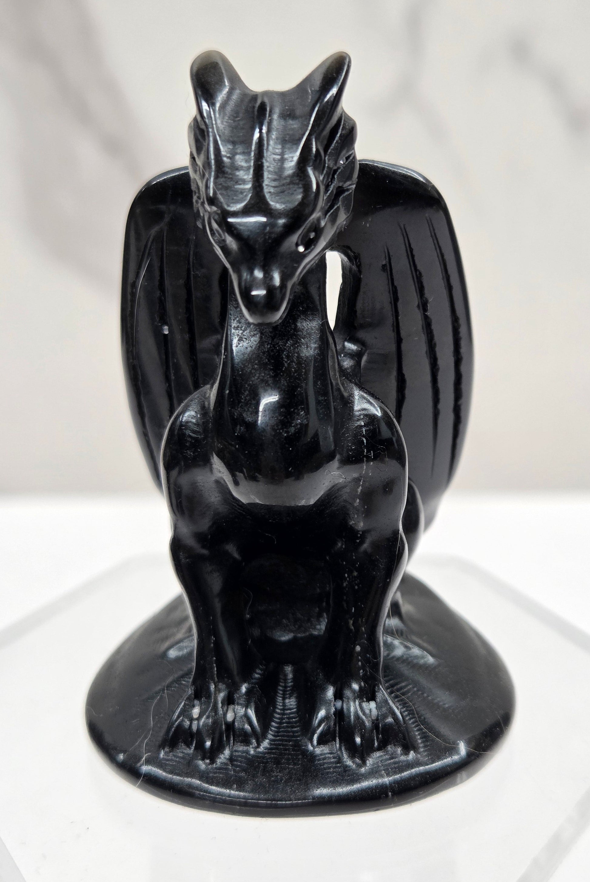 A glossy black ceramic dragon figurine mounted on a clear acrylic hexagonal display stand against a white marble-patterned background.