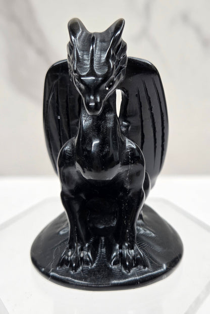 A glossy black ceramic dragon figurine mounted on a clear acrylic hexagonal display stand against a white marble-patterned background.