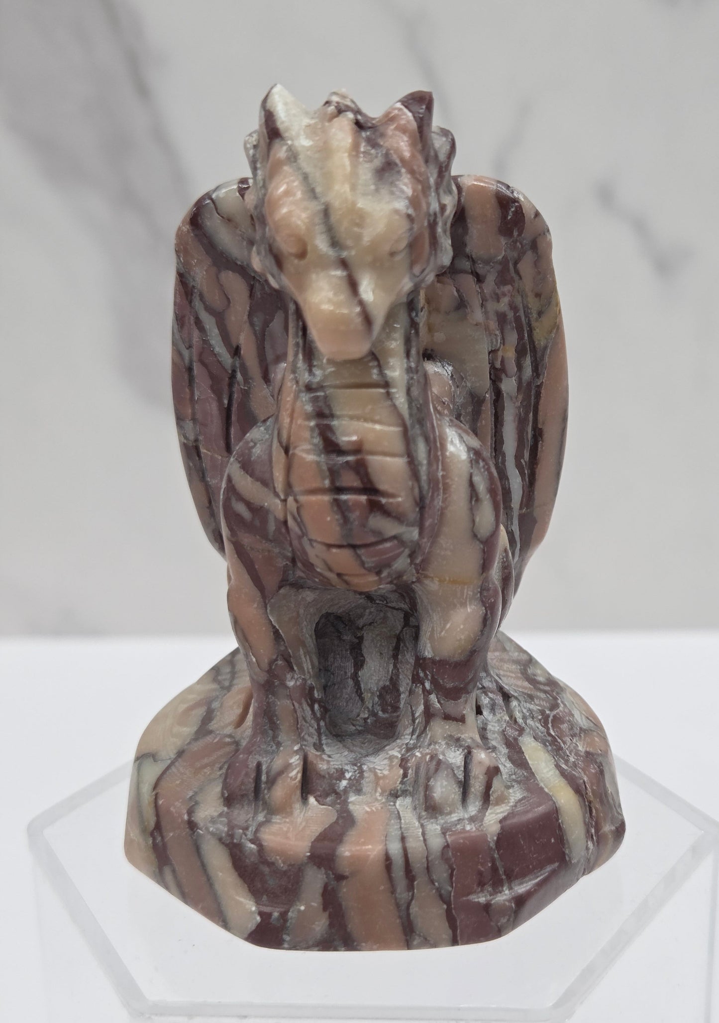 A carved stone gargoyle figurine with brown, beige, and cream marbled patterns, displayed on a clear acrylic stand against a light gray marble-patterned background.