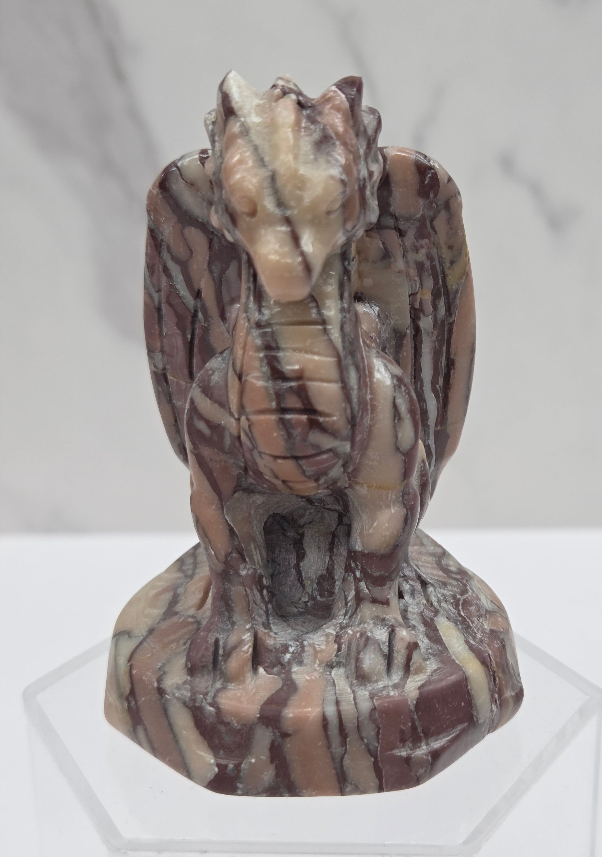 A carved stone gargoyle figurine with brown, beige, and cream marbled patterns, displayed on a clear acrylic stand against a light gray marble-patterned background.