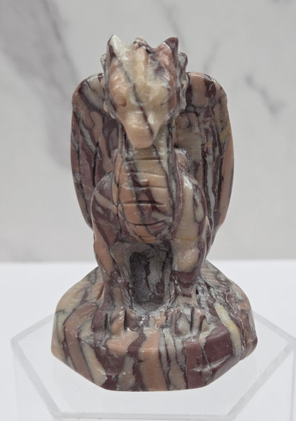 A carved stone gargoyle figurine with brown, beige, and cream marbled patterns, displayed on a clear acrylic stand against a light gray marble-patterned background.