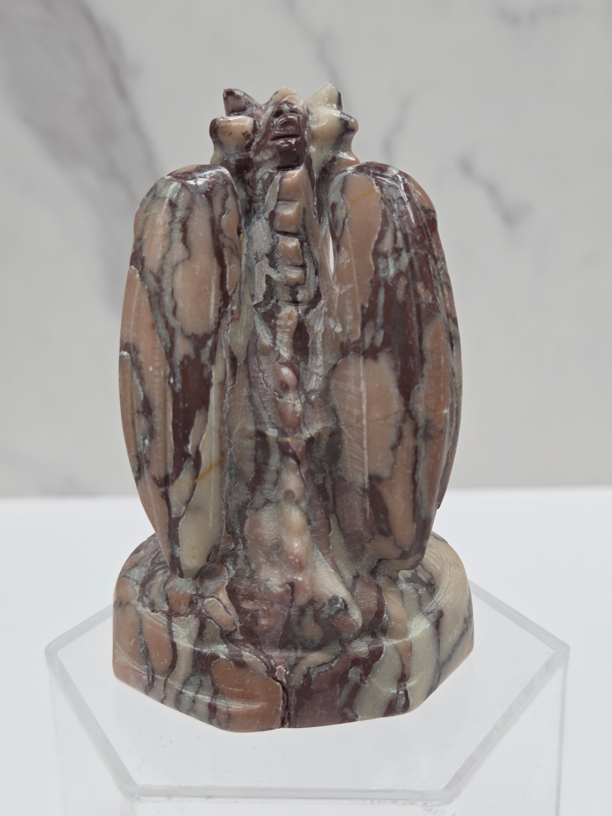 A carved stone sculpture of a winged figure, likely a gargoyle or demon, made from polished brown, beige, and gray marbled stone, displayed on a clear acrylic stand against a light gray marble-patterned background.