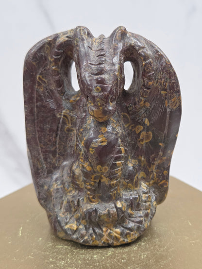 A dark brown carved dragon sculpture with intricate gold and beige inlay patterns, mounted on a gold-colored block against a white marble background.