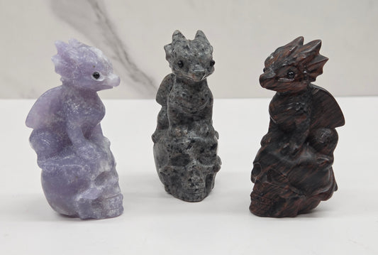 Three small dragon figurines in different stone colors, including lavender, dark gray, and dark brown, displayed on a white surface against a light gray marble-patterned background.