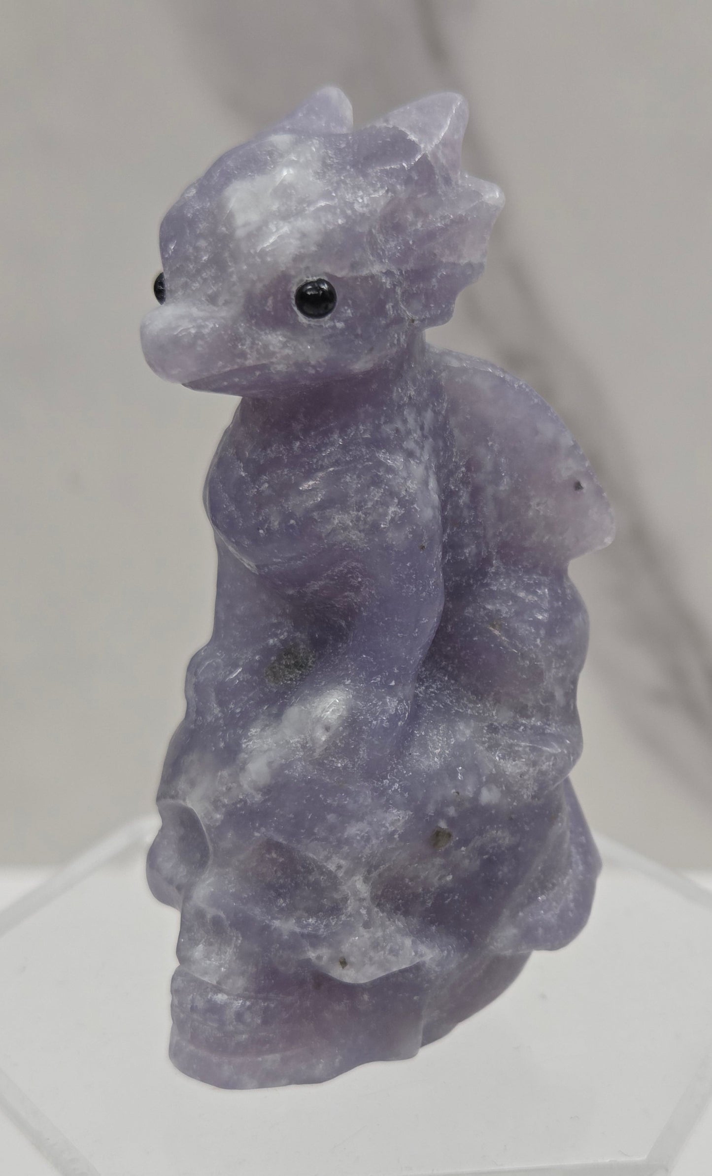 A small carved stone figurine of a dragon on a skull in a lavender-purple hue, displayed on a clear acrylic stand against a light gray marble-patterned background.