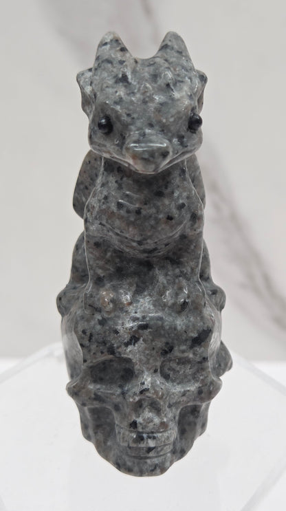 A small carved stone figurine of a dragon on a skull, made from speckled gray stone, displayed on a clear acrylic stand against a white marble-patterned background.