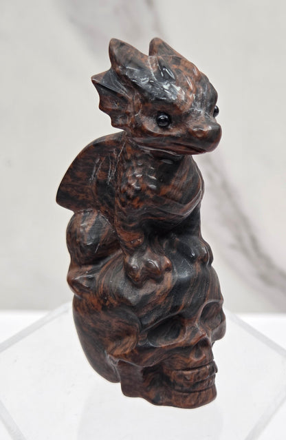 A dark carved wooden figurine dragon on a skull, displayed on a clear acrylic stand against a white marble-patterned background.