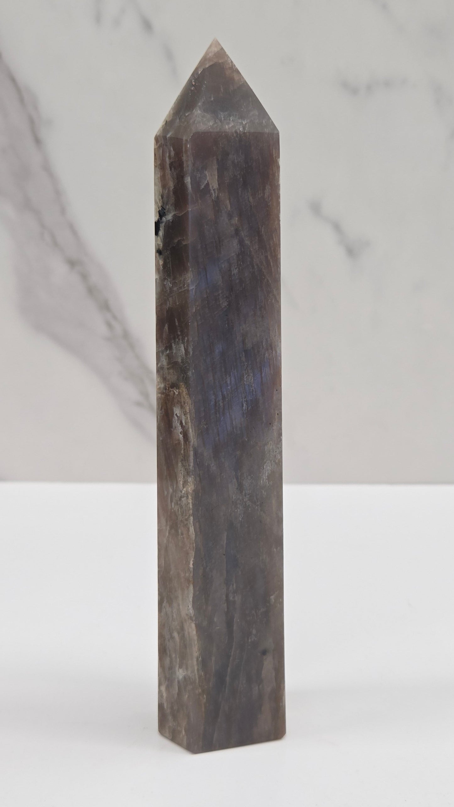 A polished dark gray crystal point wand, likely made of labradorite, resting horizontally against a white background with a marble-patterned section on the left.