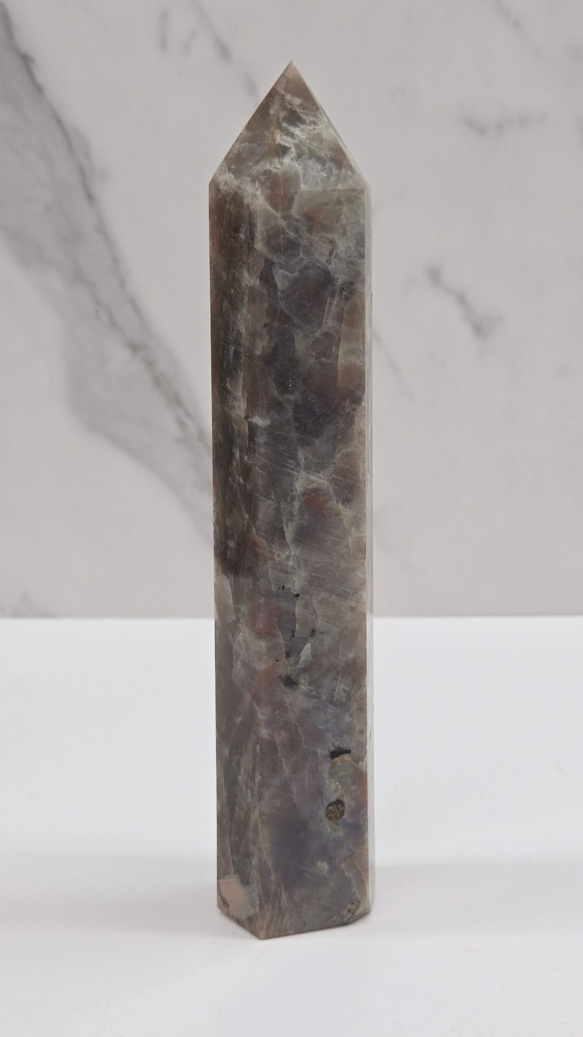 A polished rectangular crystal point with a pointed tip, made of dark gray and brown marbled stone, shown against a split background of white marble and a plain white wall.