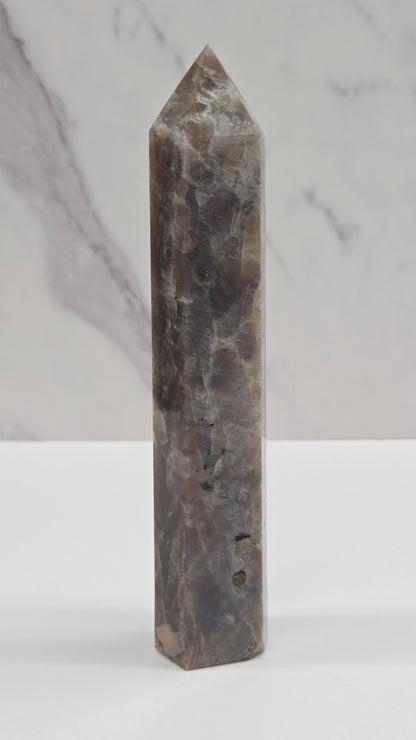 A polished rectangular crystal point with a pointed tip, made of dark gray and brown marbled stone, shown against a split background of white marble and a plain white wall.