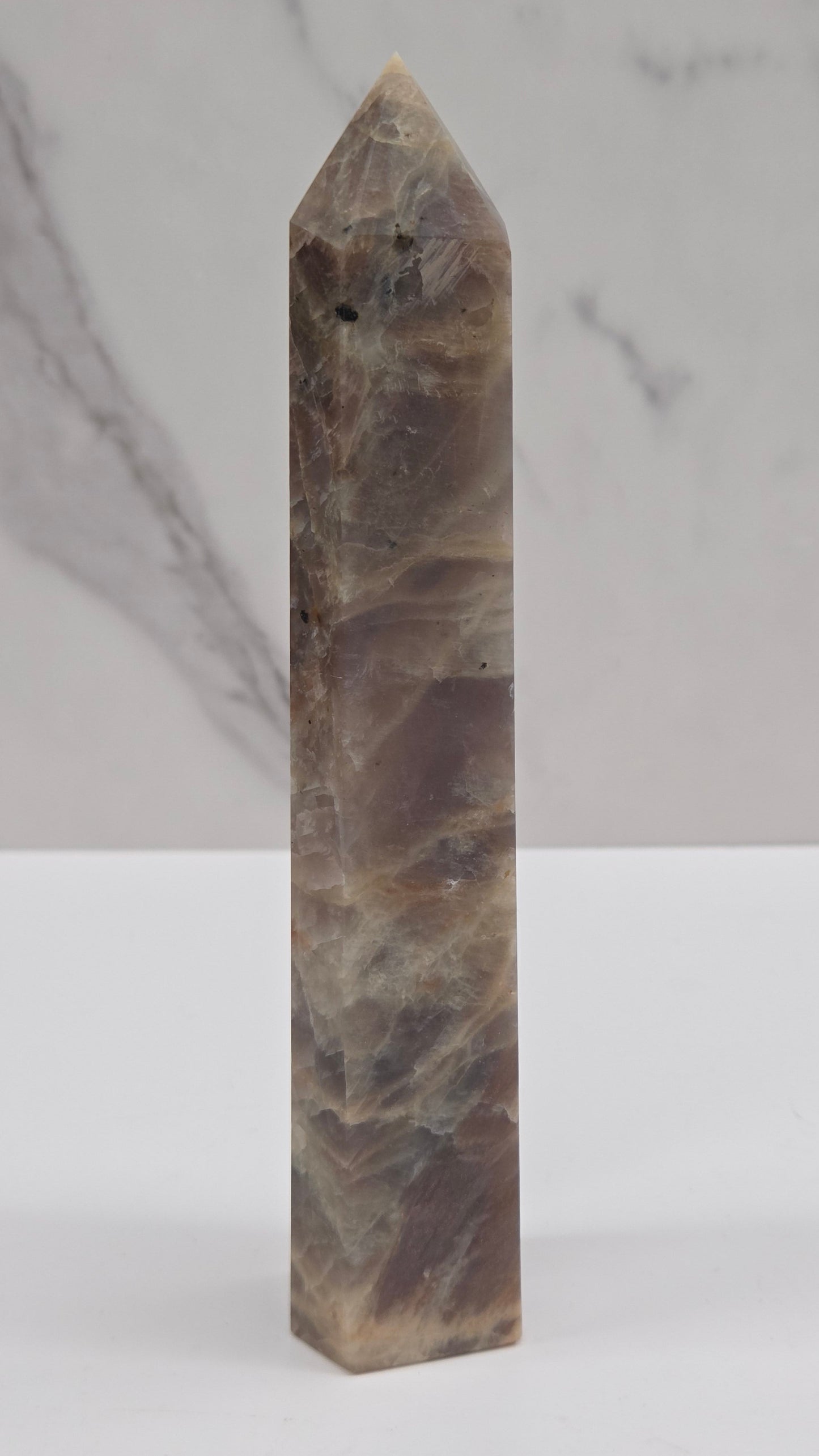 A polished natural crystal point, identified as quartz, with brown and beige marbling, displayed horizontally against a split white and white marble background.
