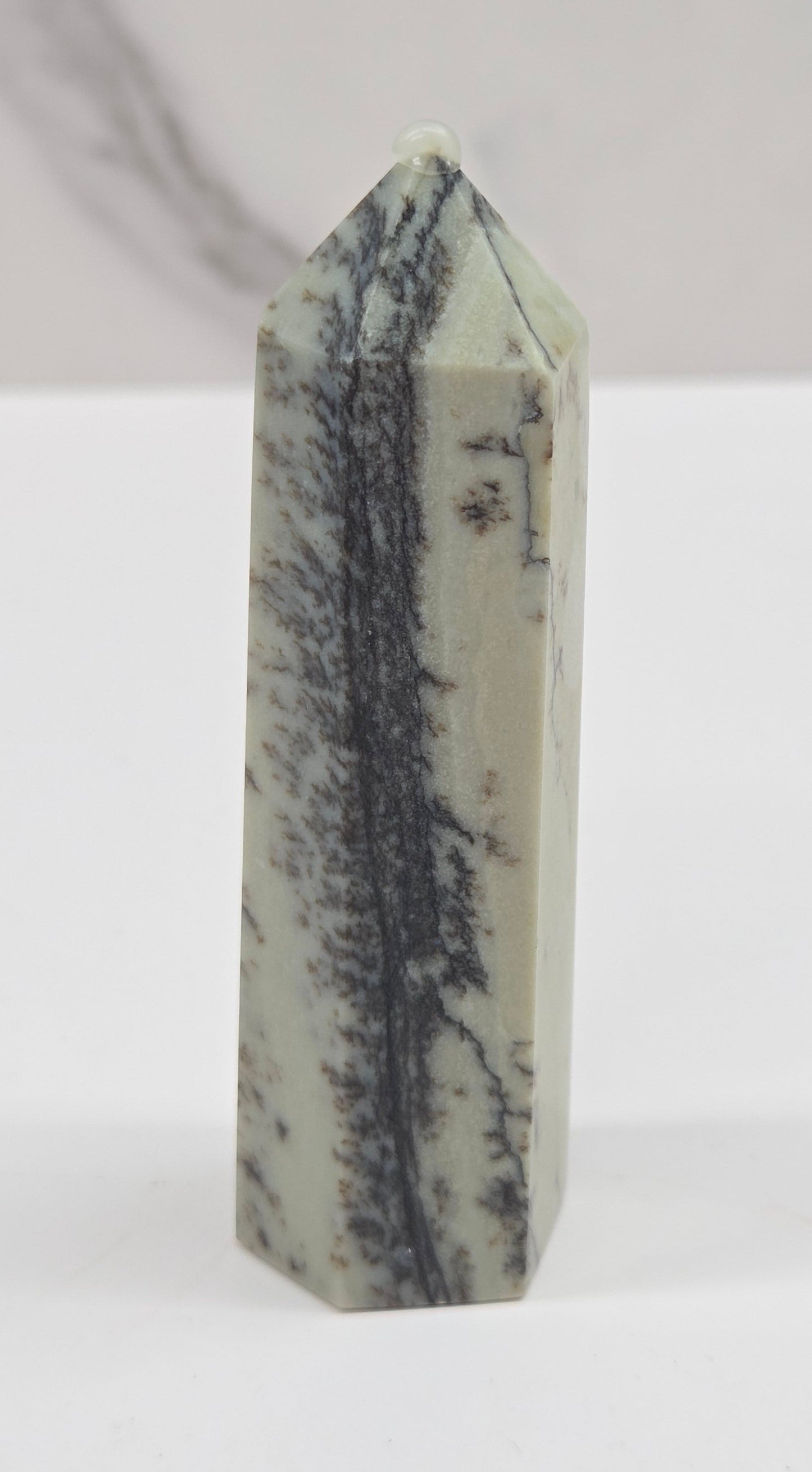 A polished pointed crystal tower, likely a quartz point, featuring a natural gray and black dendritic pattern on a pale sage green base, set against a light gray marble-patterned background.