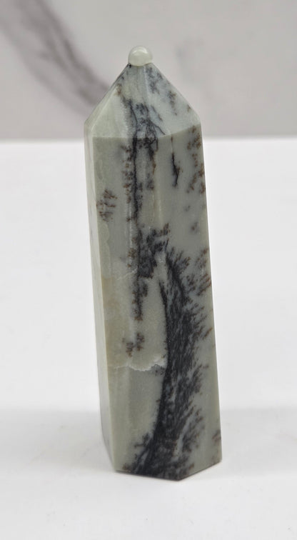 A polished natural crystal tower with a pointed top, featuring a light gray and white base with prominent black and dark gray dendritic veining, standing upright on a white surface against a light gray marble-patterned background.