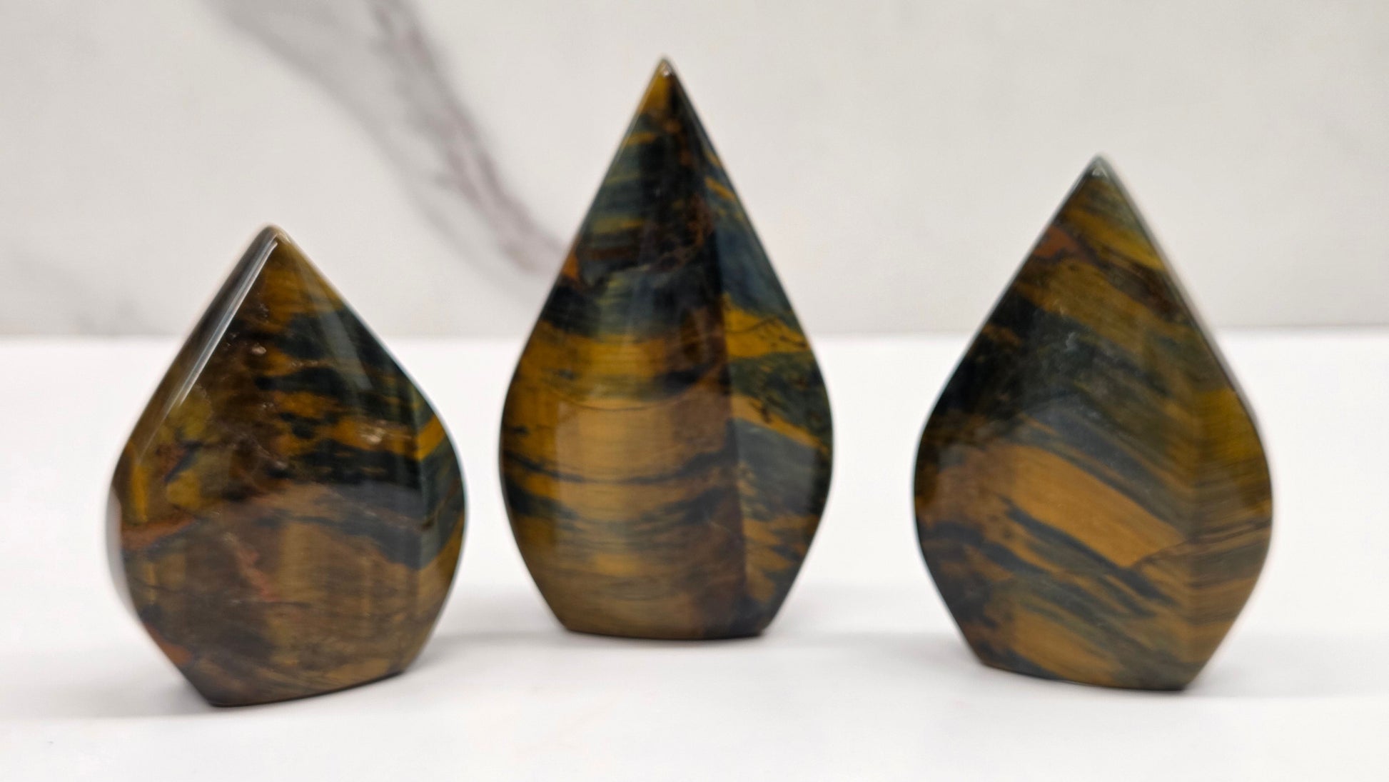 Three polished tiger's eye crystal point towers in different heights, arranged on a white surface with a light gray marble background.