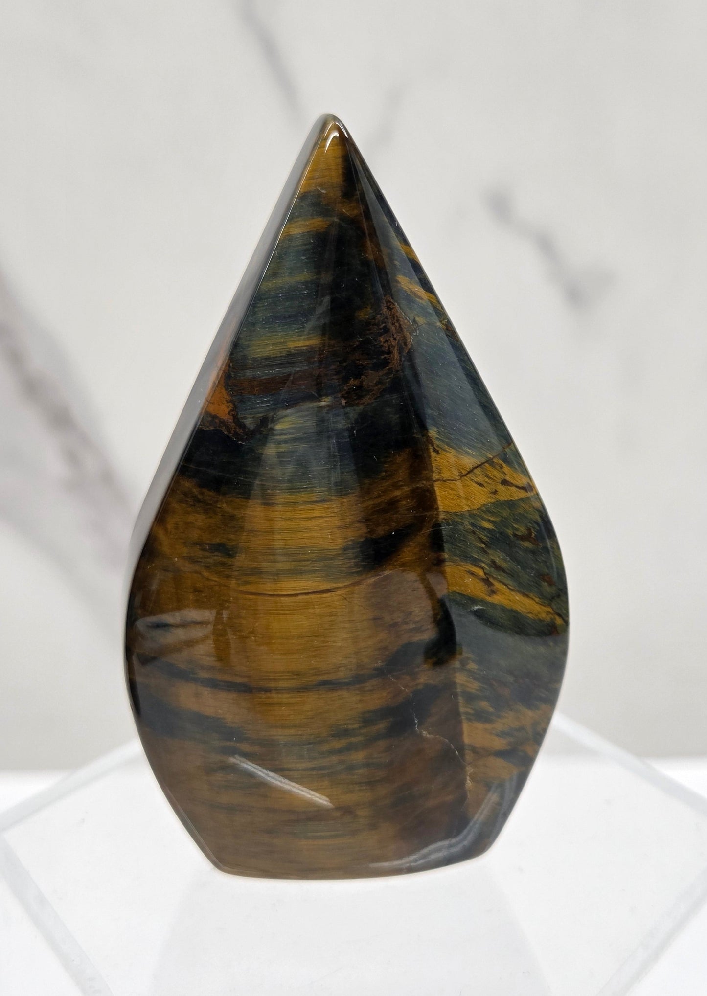 A polished tiger's eye crystal point sculpture displayed on a clear acrylic stand against a white marble background.