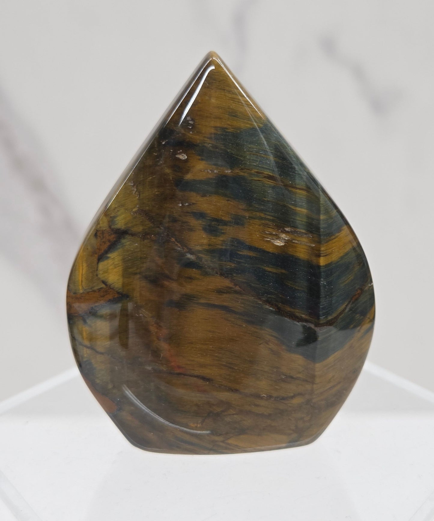 A polished tiger's eye gemstone cabochon with natural brown and green banded patterns, displayed on a clear acrylic stand against a white marble background.