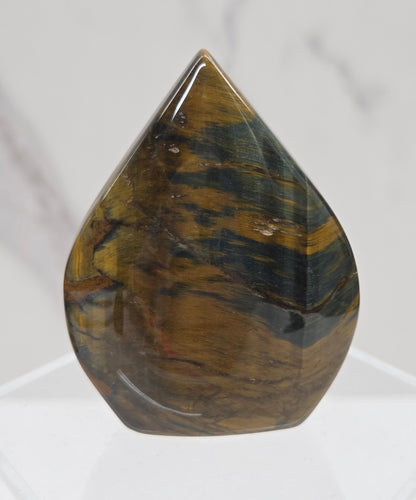 A polished tiger's eye gemstone cabochon with natural brown and green banded patterns, displayed on a clear acrylic stand against a white marble background.